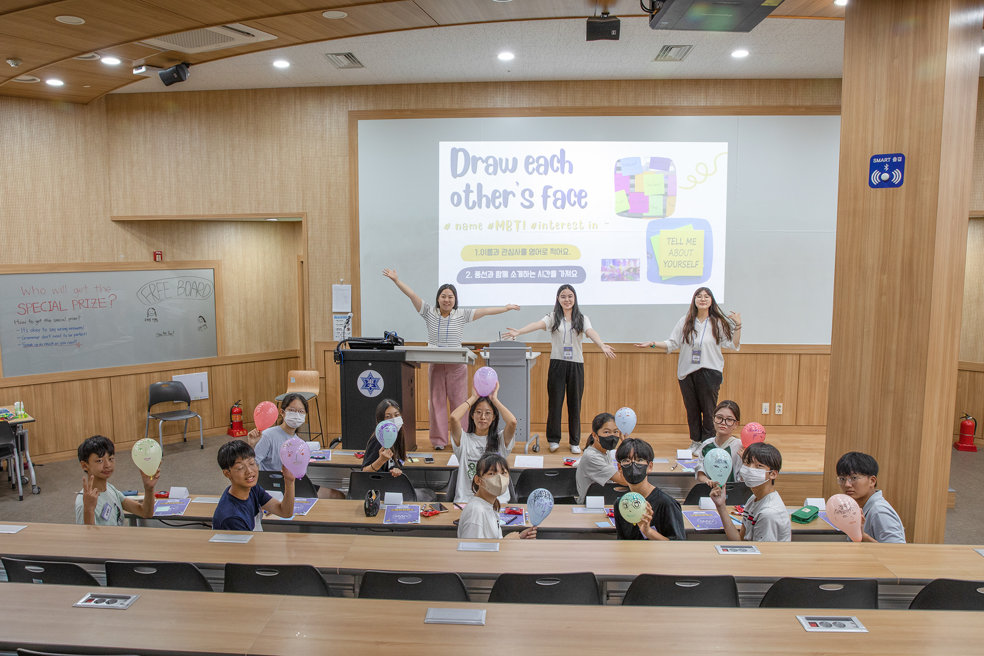 작은 강당에서 남녀 아이들과 선생님이 카메라를 보며 사진 찍는 자세를 취하고 있는 사진