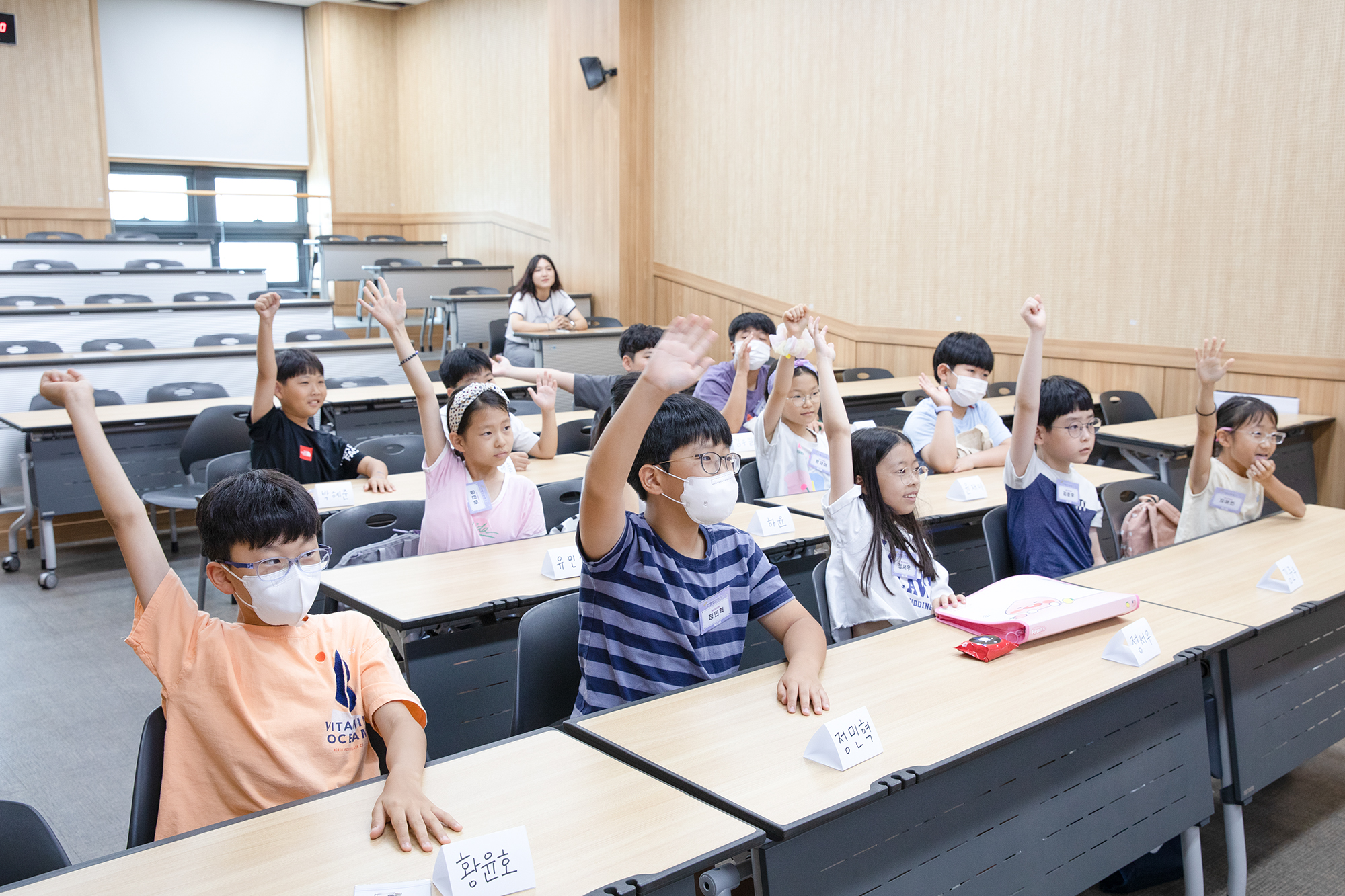 남녀 어린이들이 발표를 위해 한쪽 손을 번쩍 들고 선생님을 바라보고 있는 사진
